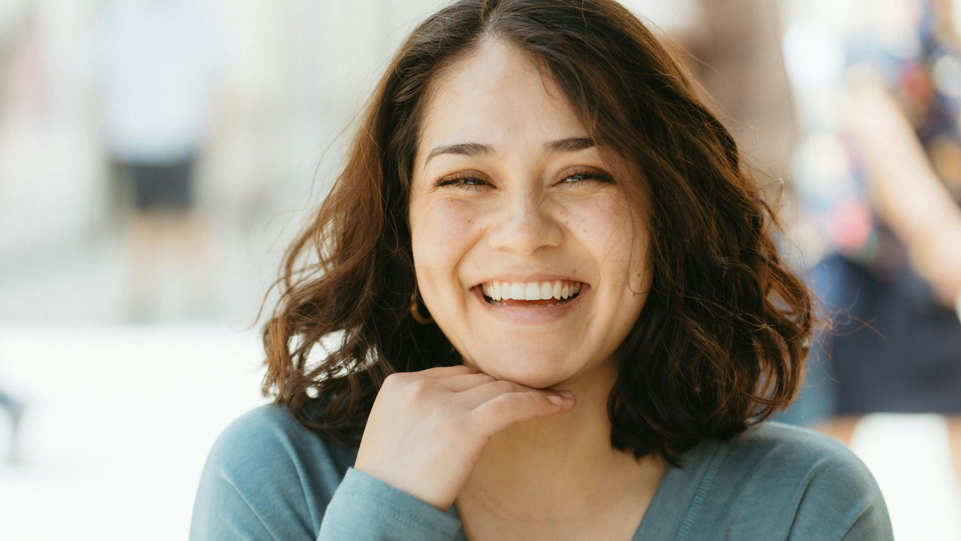 A young woman with curly brown hair laughing warmly, resting her chin on her hand, wearing a light blue top.